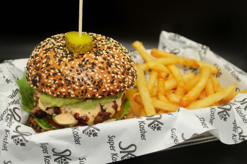 Hamburguesa con pan de ajonjolí, pepinillo y papas fritas servida en papel con el logo de Super Kosher