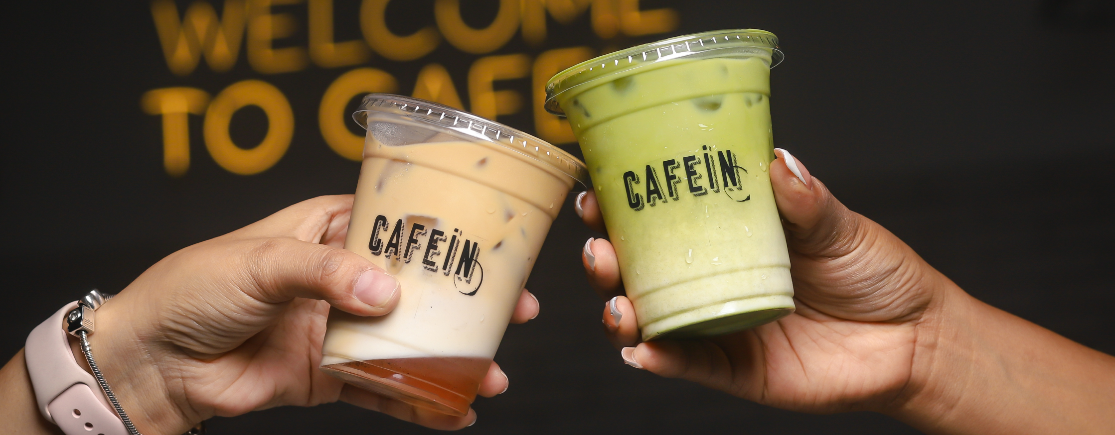 Dos manos brindan con un iced coffee y un matcha frío en vasos de Cafein frente al letrero Welcome to Cafein