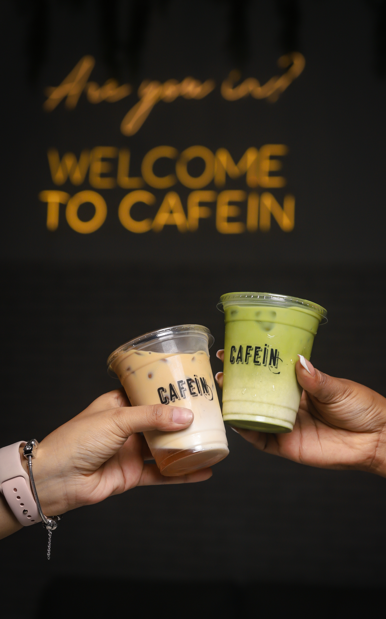 Dos vasos de Cafein frente al letrero luminoso Welcome to Cafein