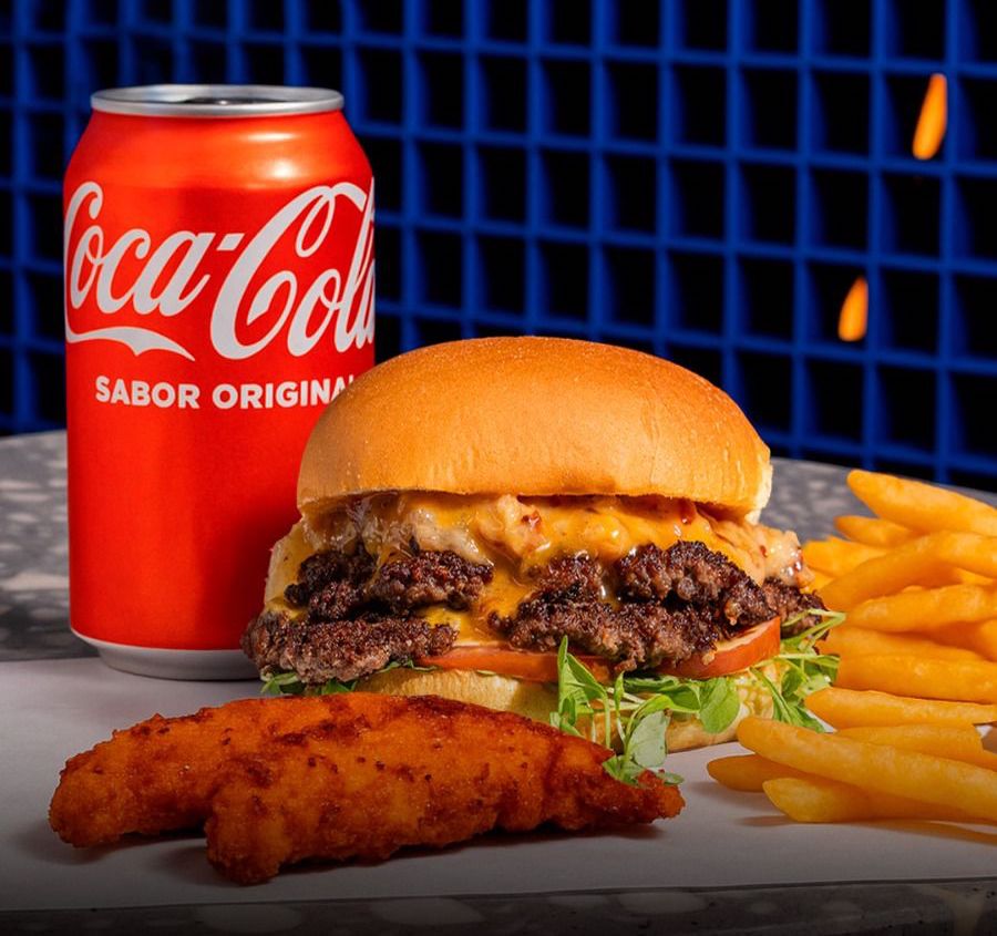 Smash burger doble con queso de Naked Lukas junto a fries, un chicken tender y una Coca-Cola sobre fondo de azulejos azules