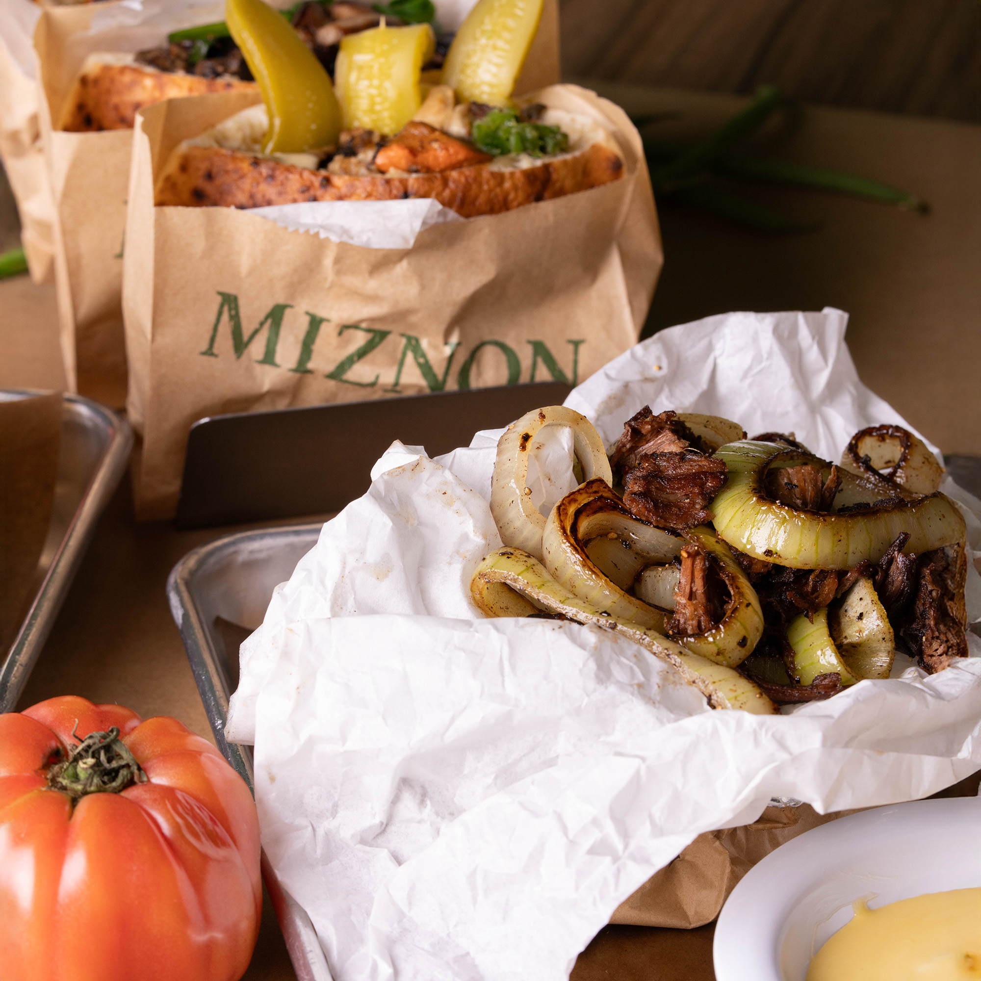 Pitas en bolsas kraft de Miznon junto a una porción de carne y cebolla a la plancha envuelta en papel y un tomate grande