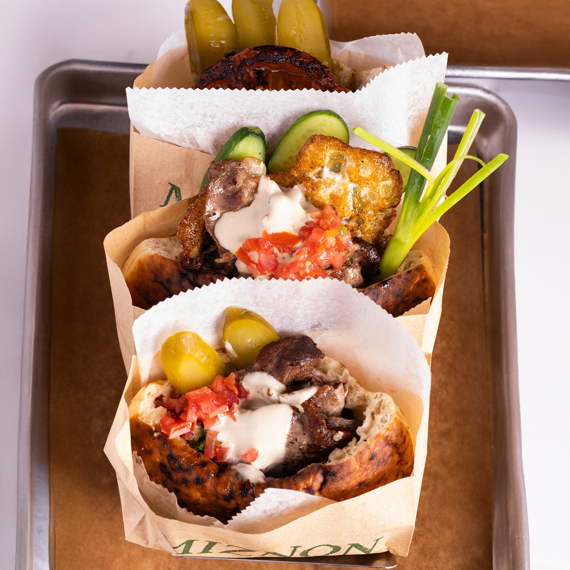 Tres pitas de Miznon en bolsas de papel kraft, rellenas de carne, tahini, tomate picado, pepinillos y cebollín sobre una bandeja de metal