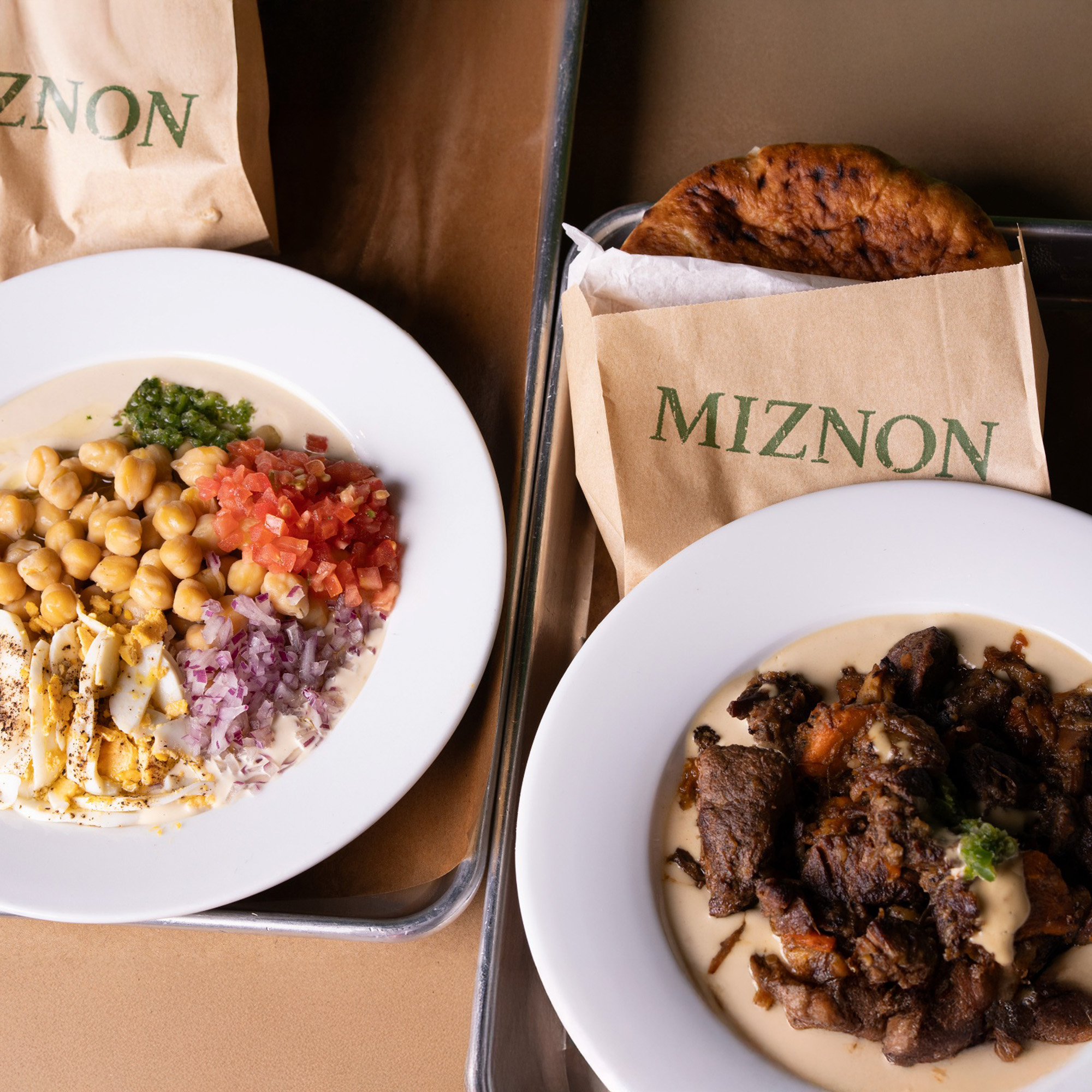 Plato de garbanzos con tomate y cebolla morada picados, plato de carne guisada sobre crema y una pita en bolsa de Miznon