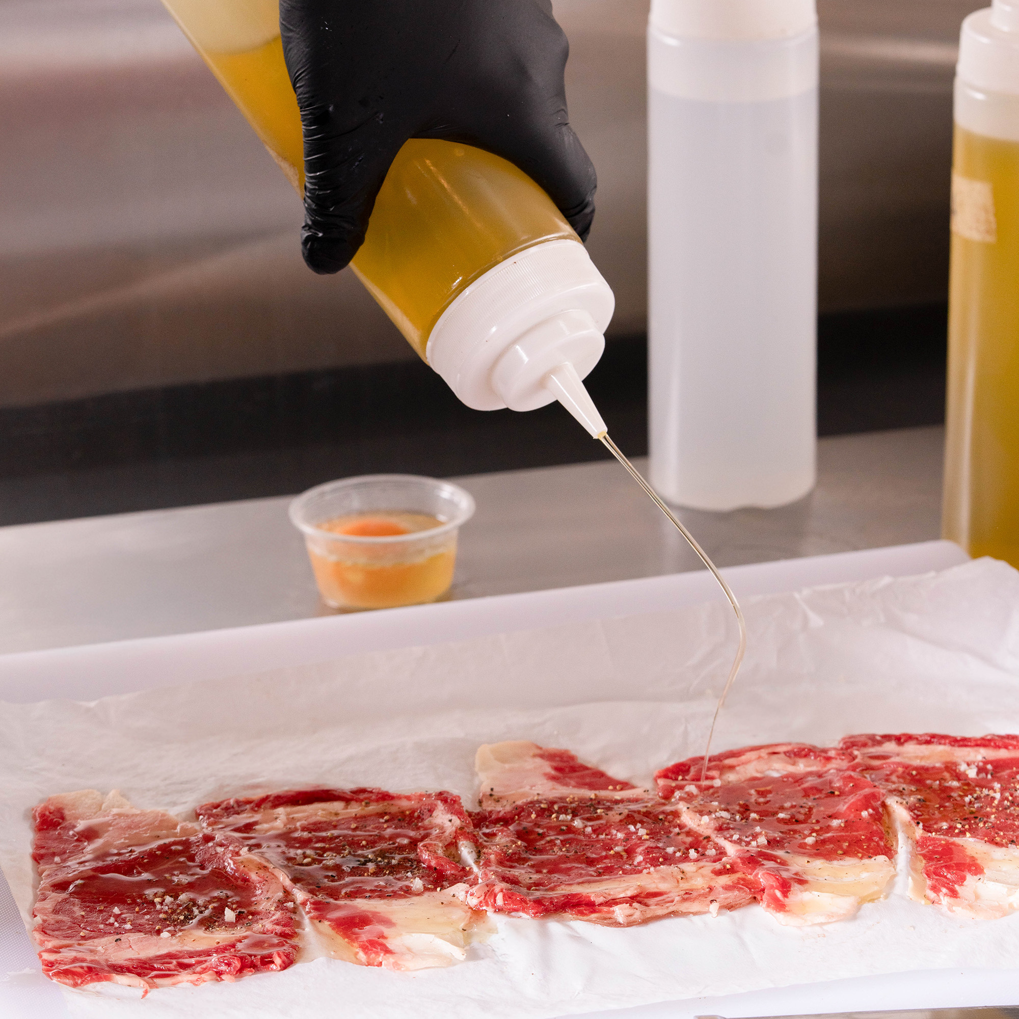 Lonjas de carne cruda marmoleada sobre papel mientras una mano enguantada sirve aceite