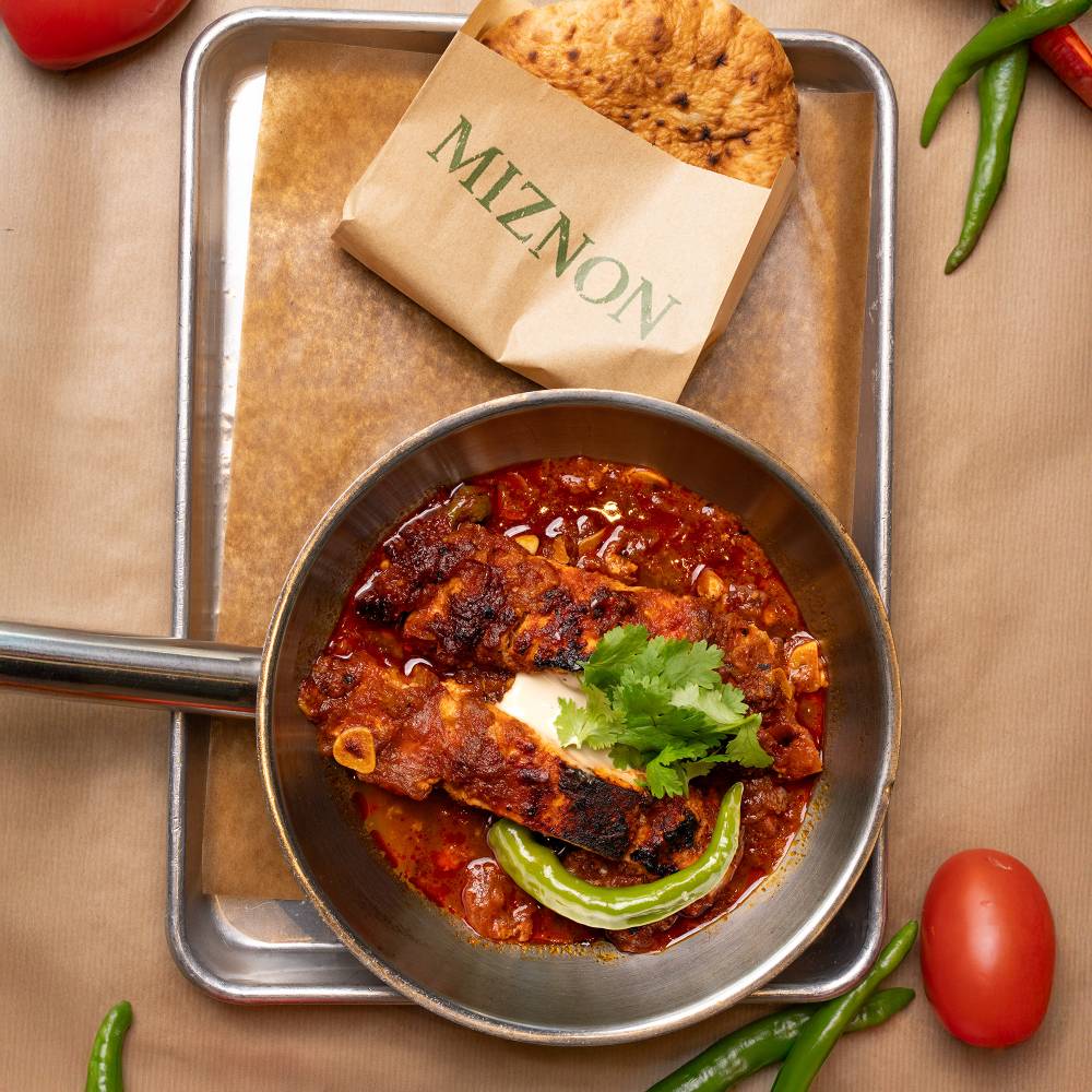Sartén con salsa de tomate, cilantro y ají verde junto a una pita en bolsa de papel de Miznon