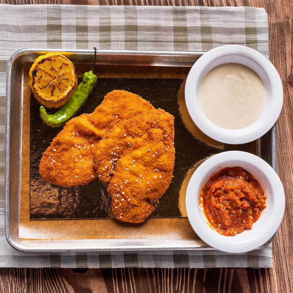 Milanesa dorada en bandeja con limón asado, ají verde, tahini y salsa roja picante
