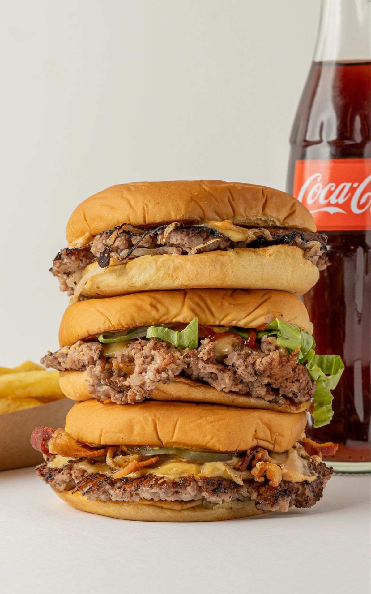 Tres smash burgers de Miss Patty Bam Bam apiladas junto a una botella de Coca-Cola