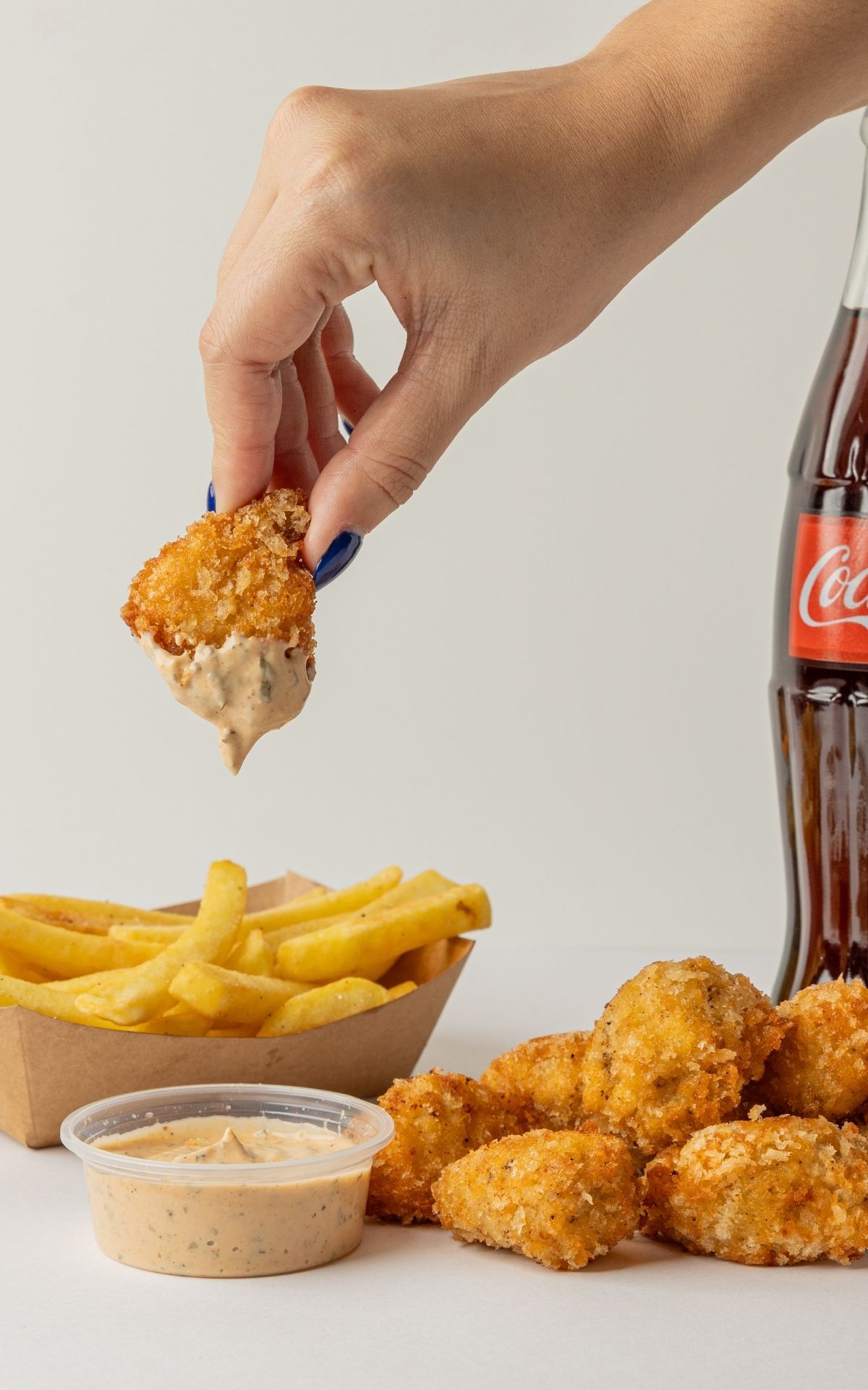 Mano mojando un chicken bite en salsa, con papas fritas y Coca-Cola al fondo